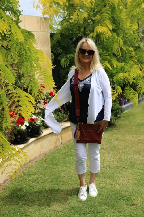 Blond woman carrying a crossbody bag, standing in a garden, wearing white and navy blue clothes.