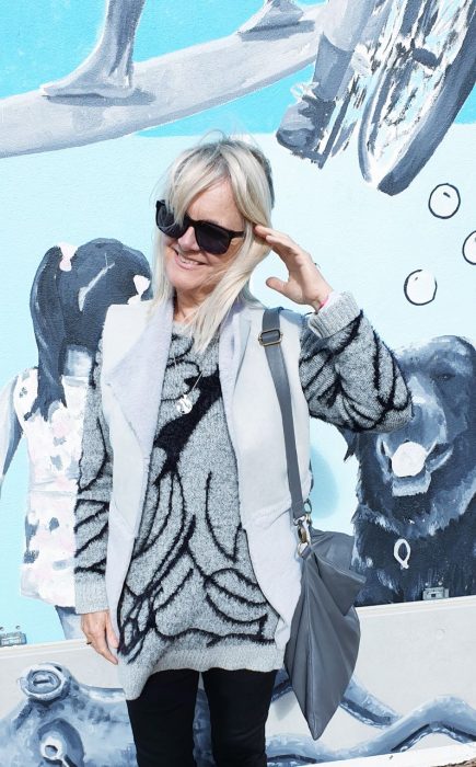 Blonde woman in dark glasses wearing a grey faux fur vest and a black and grey sweater standing against a mural with a dog. She's carrying a grey shoulder bag.