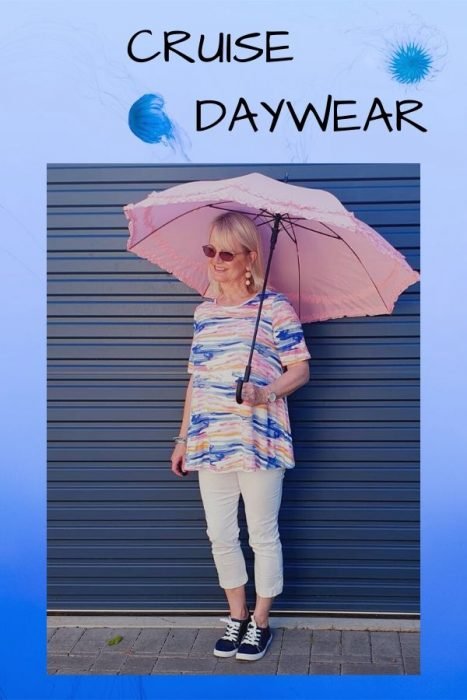 Blond woman carrying pink umbrella wearing stripey pink and blue top over white capri pants with black and white sneakers on her feet. Clothes suitable for Cruise daywear