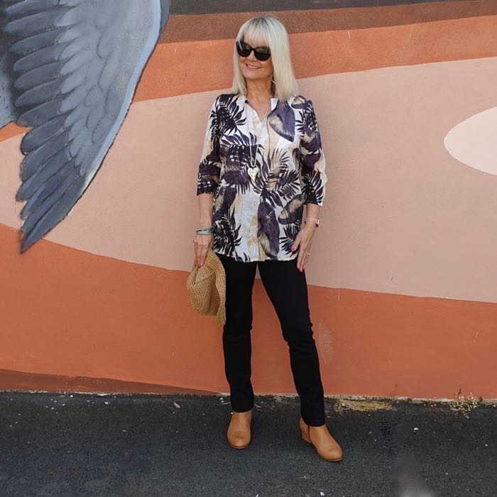 Blonde woman in black trousers and a caramel, white and black blouse. She's wearing tan ankle boots and carrying a straw hat. Illustrating how to dress well if you are short.