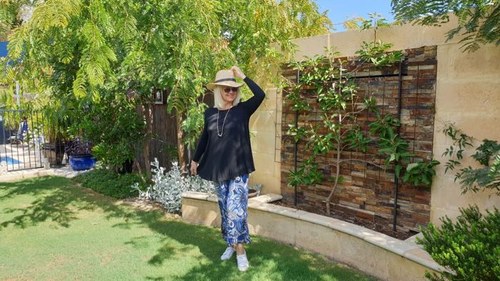 Woman standing in a garden. She is wearing a straw hat, baggy black top and blue and white baggy pants illustrating how the right dimensions are necessary if you are a short woman.