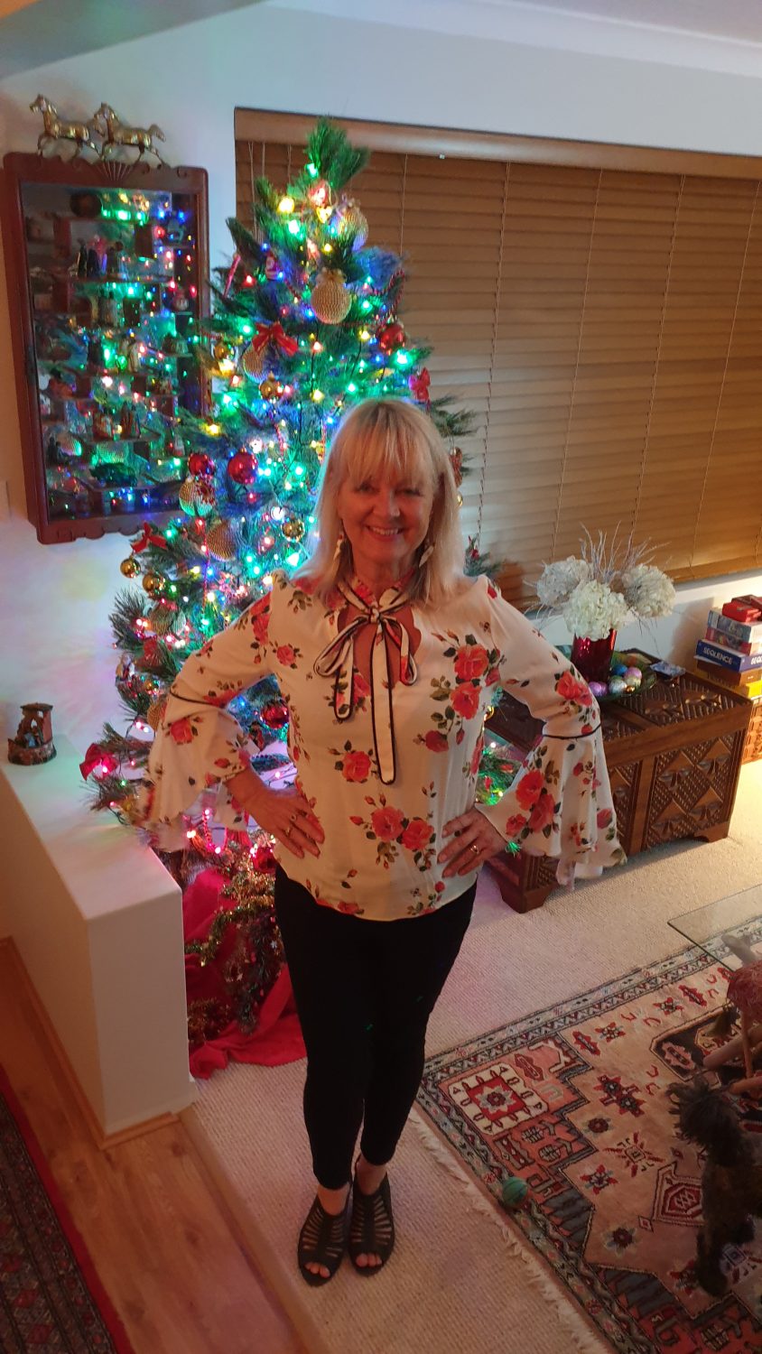 Woman in flowery flared sleeve top and black slimline pants standing in front of a Christmas tree, showing how skinny jeans can make your legs look shorter.