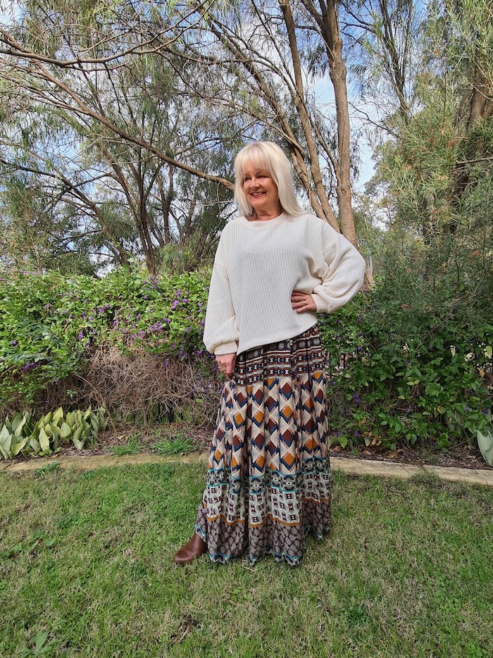 Blond haired woman wearing dark sunglasses and an ivory colored sweater over a paisley print maxi dress.