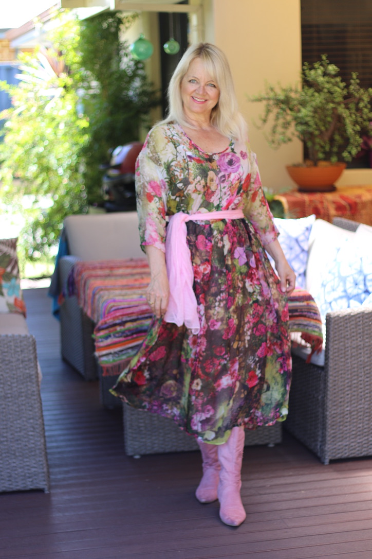 Blong haired woman wearing a silk dress that has a floral pattern and a pink scarf tied around the waist.