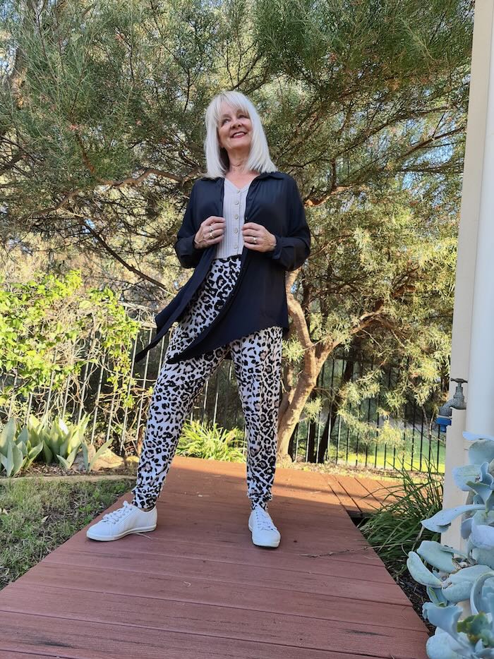 Blond woman over the age of 50 years old wearing sensational animal print pants and a camel top with a black technical shirt worn as a jacket. On her feet she is wearing white sneakers.