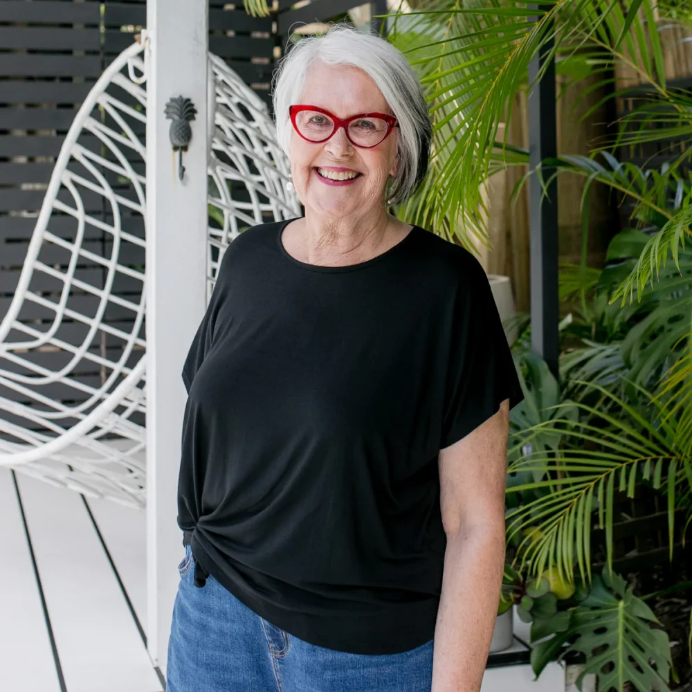 Grey haired woman wearing red rimmed glasses and a black tee shirt over blue denim jeans.