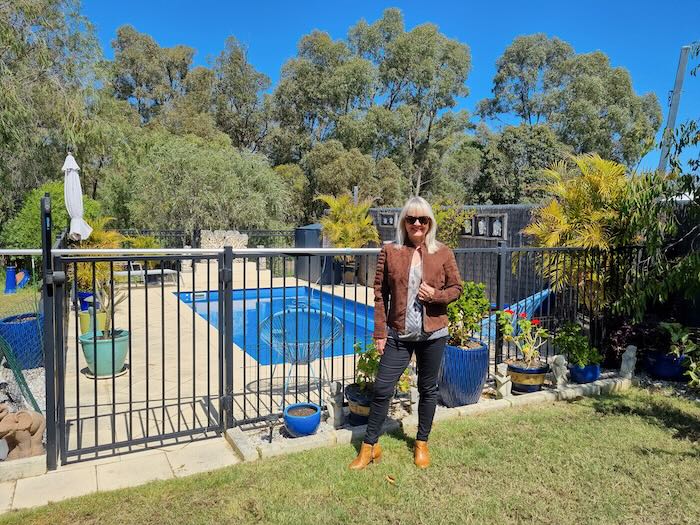 Woman standing by a swimming pool. She is wearing a suede leather jacket, black pants and chestnut coloured ankle boots.