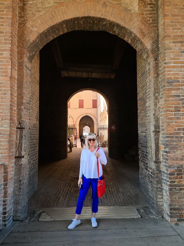 Woman standing in an archway. She is wearing a long sleeved white T shirt and blue pants. On her feet are white sneakers and she is carrying a red handbag.