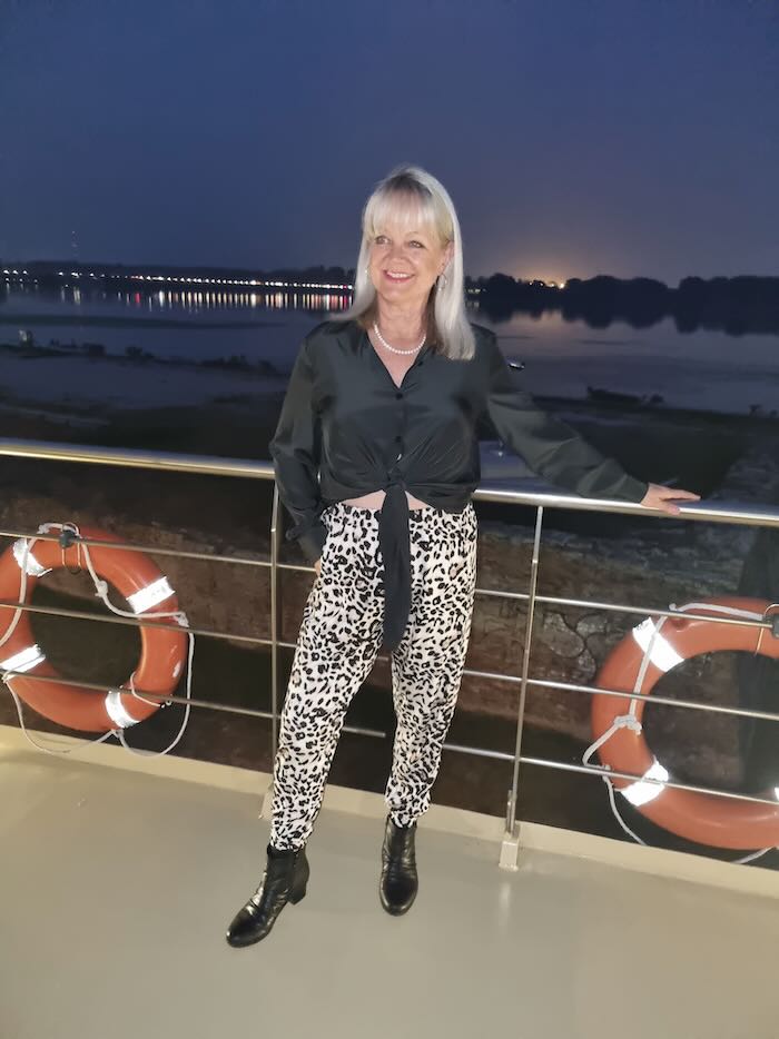 Woman standing on a boat by the railings. She is wearing a black shirt and leopard print pants with black boots.