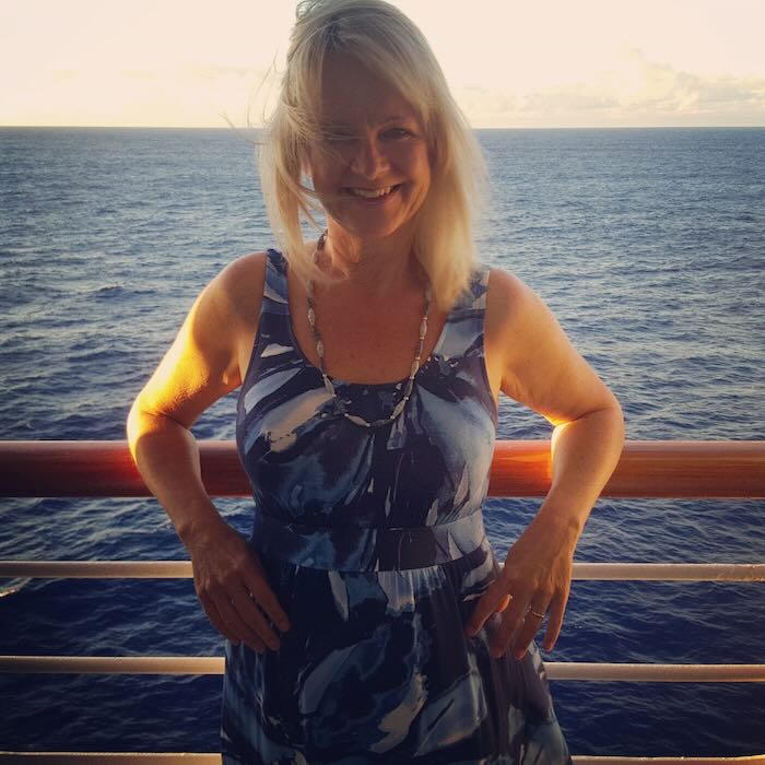 Woman wearing a blue dress with a fitted bodice. She is standing by the railings on a cruise ship out at sea. The dress is a good example of a style to suit pear shaped women.