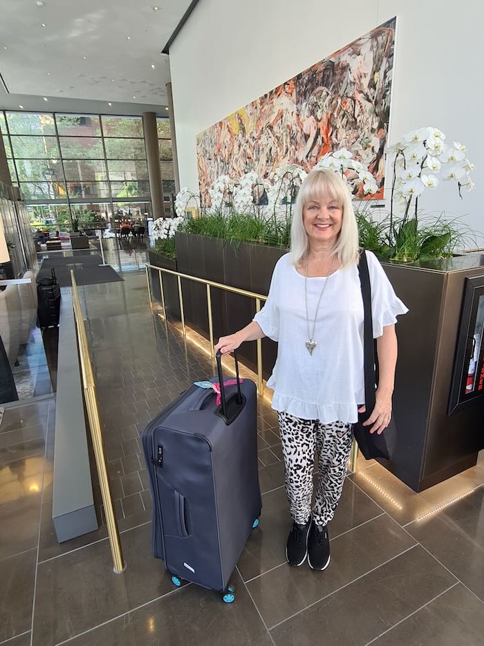 Blond woman smiling confidently in a hotel lobby. She is wheeling a suitcase. She is wearing casual clothes suitable to pack for an Alaska cruise in May.