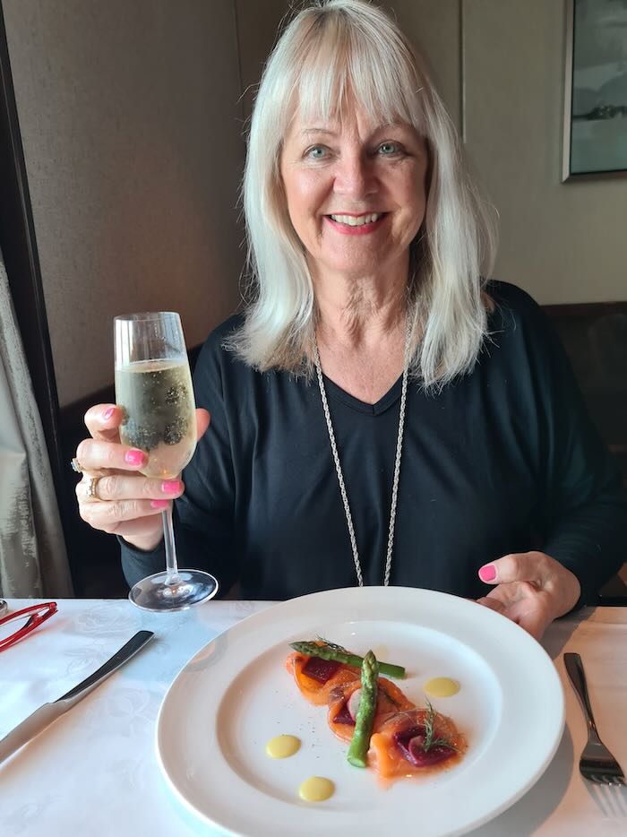 Woman on a cruise ship sailing to Alaska, dining, saying cheers with a glass of champagne. She is wearing a black blouse.