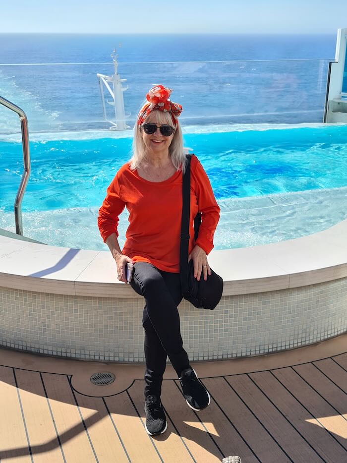 Woman sitting by swimming pool on cruise ship during a cruise to Alaska. She is wearing an orange Tee shirt and black pants. She has a bow tie in her hair.