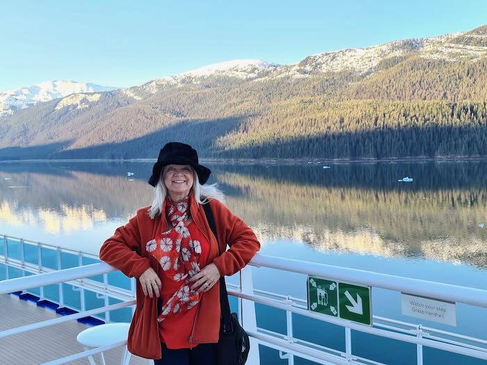 Woman standing on a cruise ship beneath snowy mountains. She is wearing a black hat, orange jacket, orange Tee and flowery silk scarf, all suitable garments to wear for a cruise to Alaska.