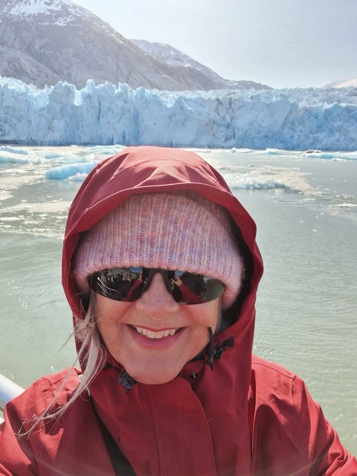 When thinking about packing for Alaska Cruise in May, layering is essential as seen here on this woman standing near a glacier. She is wearing a waterproof jacket and a pink beannie, with dark sunglasses.