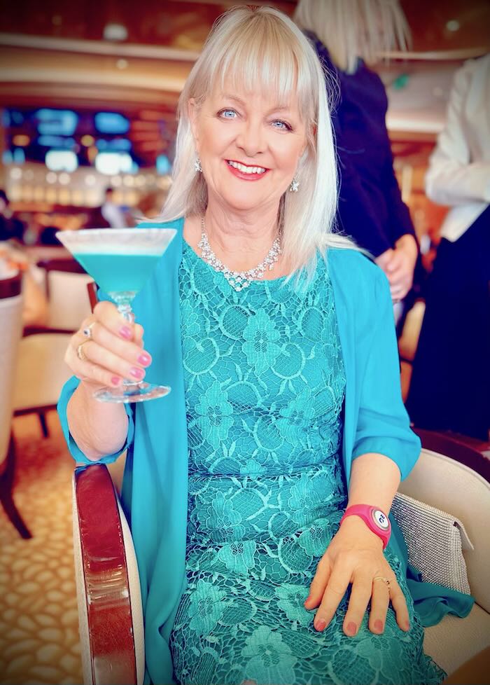 Blond woman wearing a turqoise blue cocktail dress. She is holding a blue cocktail. She is showing an item of clothing suitable to pack for a formal night on a cruise in the post what to wear for an Alsaka cruise in May.