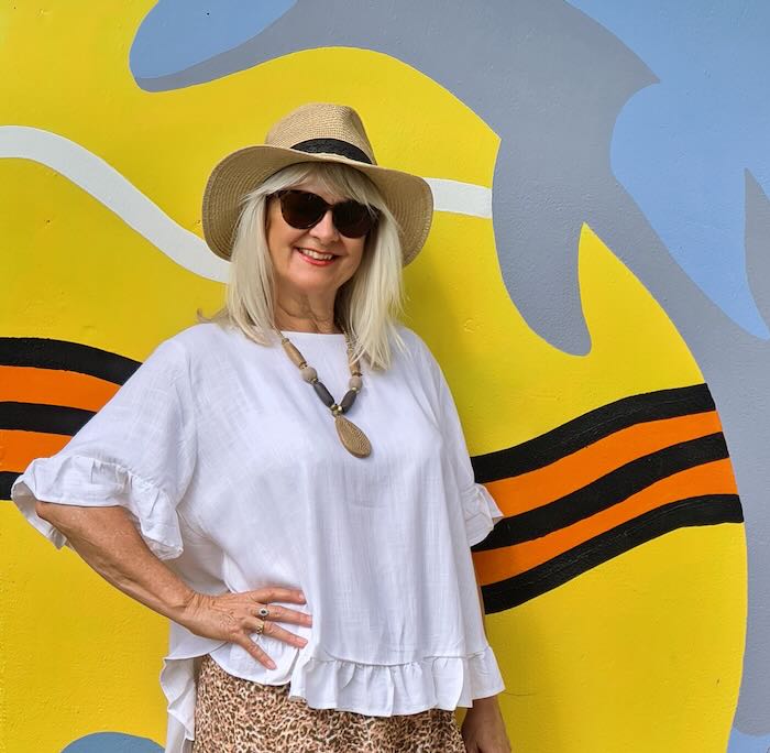 Blond haired woman wearing a straw hat, dark sunglasses and boho chic outfit. White floaty blouse and animal print wide leg pants.