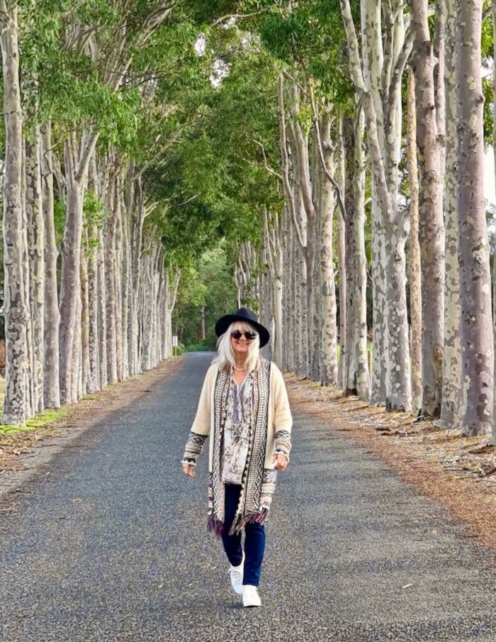 Blond haired woman walking down an avenue of tall trees. She is wearing a boho chic outfit consisting of black hat, dark sunnies, long cardigan with tassles, floaty floral blouse and white sneakers.