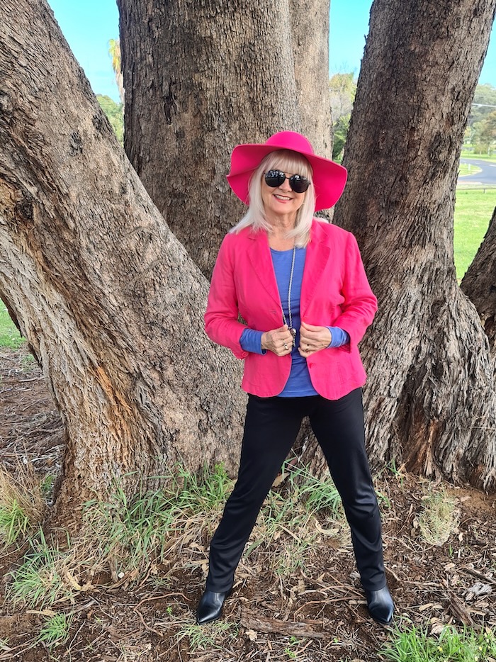 Blond lady in pink hat standing by a tree. She's wearing a pink jacket, blue top and black pants.