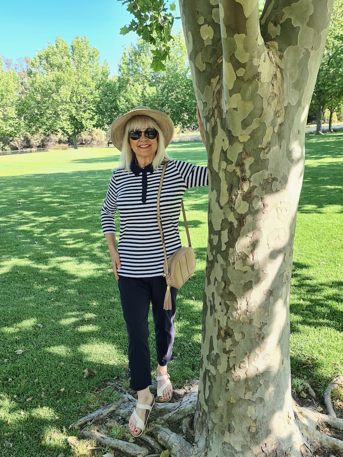 Woman wearing a hat and a stripey top with blue pants and a beige handbag. The clothes she is wearing are suitable to include in a basic wardrobe for women over 60.