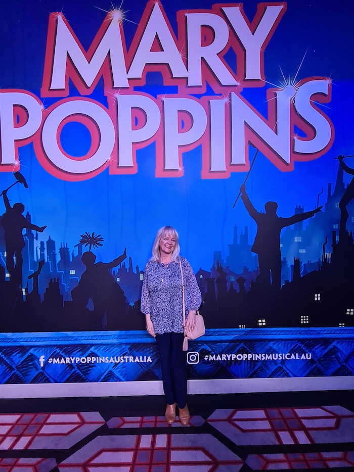 Woman standing in front of a Mary Poppins the musical poster. She is wearing going out clothes suitable for a woman over 60.