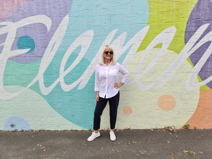 Woman wearing a white blouse and navy pants standing in front of a street mural.