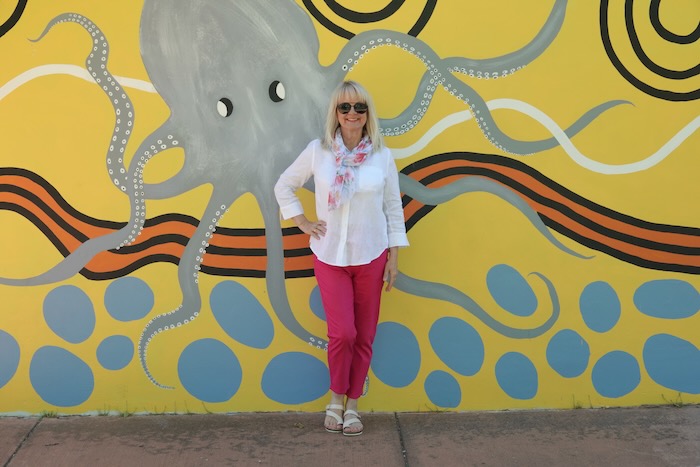 Woman wearing a white bloues, pink pants and a pretty scarf. She is standing in front of an octopus street mural.