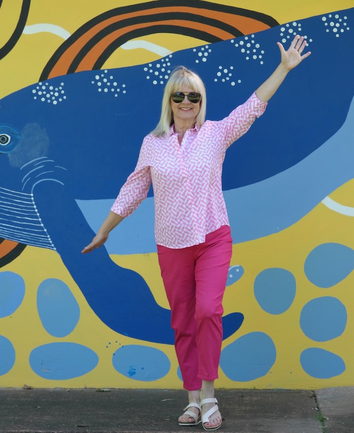 Mature age model smiling, standing in front of a street mural. She is wearing pink pants and a pink and white blouse with flat sandals. She is showing clothes that are suitable for creating a basic wardrobe over 60.