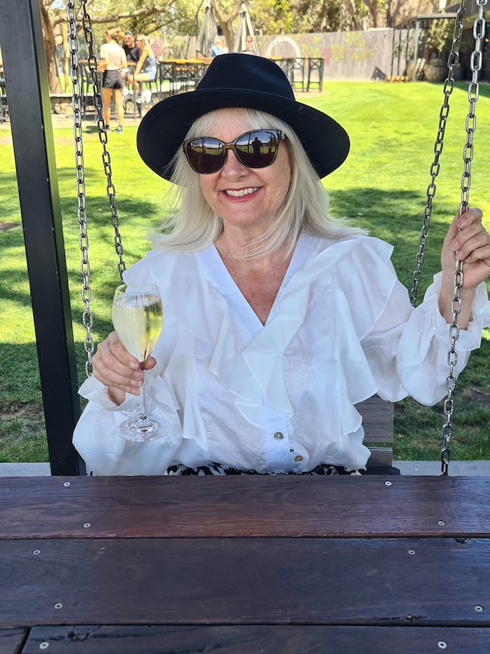 Jo wearing a black hat suitable to go with a boho outfit if you're over 50. She's also wearing a boho inspired white blouse and holding a glass of bubbles.