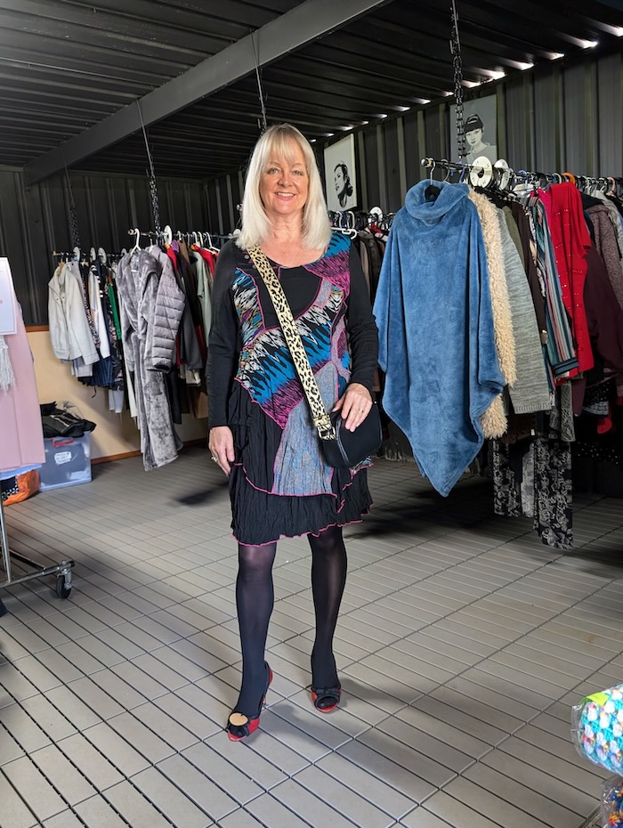 Woman helping to raise money for charity by buying clothes from an Op Shop. She is wearing a multi-colored black dress and black handbag.