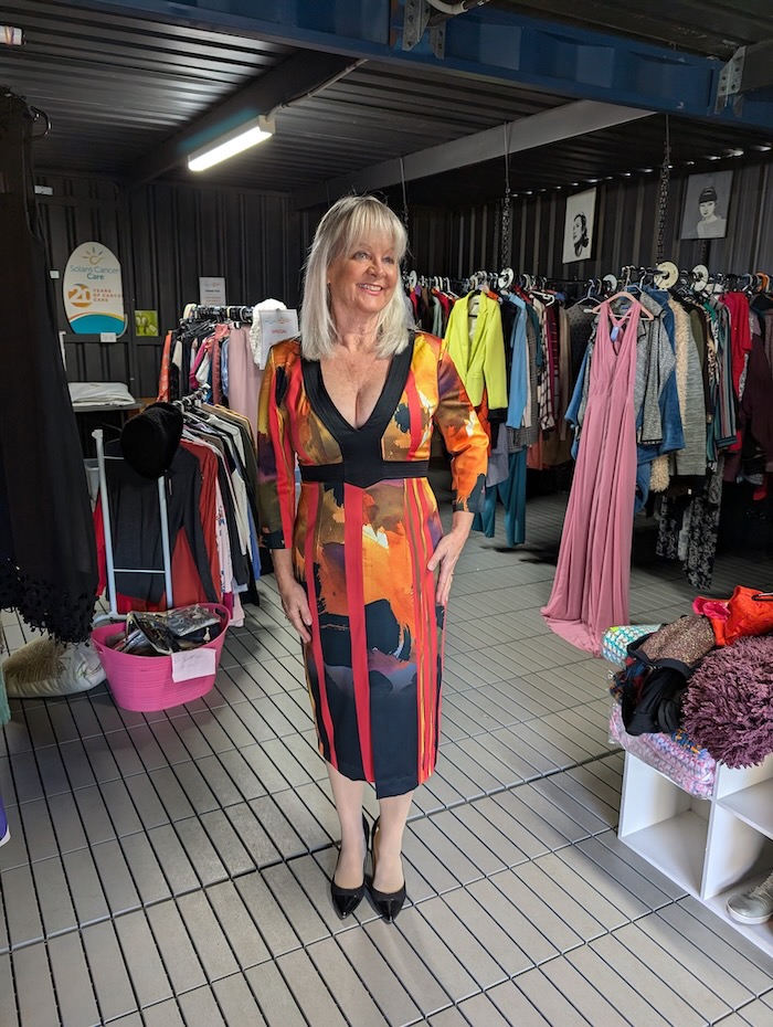 Woman standing in a thrift store. She is wearing a multicolored dress that she has bought in aid of charity.