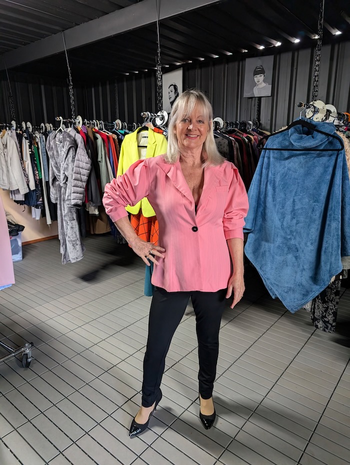 Woman standing in an Op Shop modelling a pink jacket and black pants.