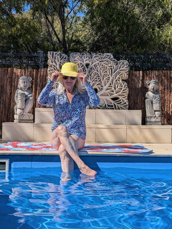 Woman sitting on a towel by a swimming pool. She is wearing sun protective clothing and a yellow sun hat.