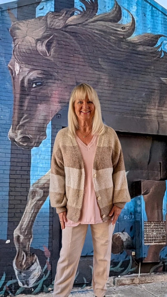 Woman in front of a mural wearing a pink sweater, a beige striped cardigan and corduroy pants. She is dressed to look a little bit Boho.