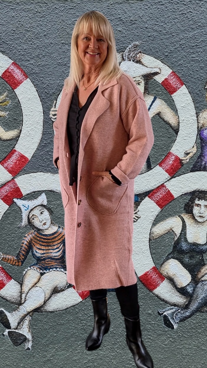 Blond woman in front of a mural wearing a long pink cardigan, black pants and black ankle boots. 